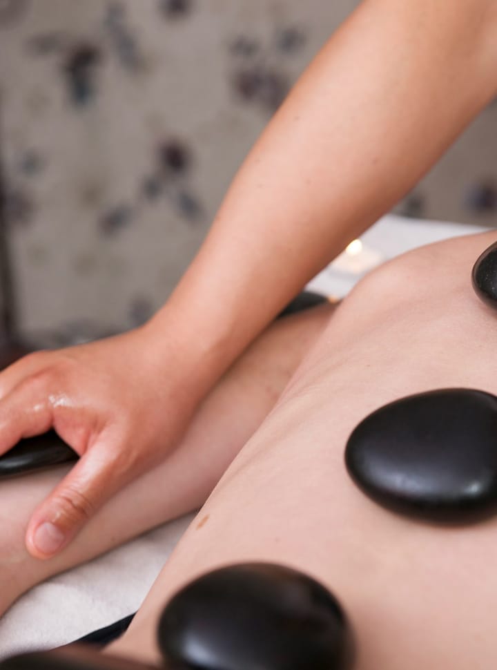 Femme couchée sur le ventre avec des pierres noires posées sur le dos, une autre personne la masse.